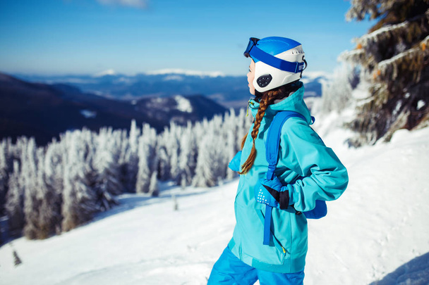 A young girl enjoys beauty of winter landscape. A beautiful girl in winter clothes, a blue helmet and jacket is having a great time in the mountains. Concept of travel, leisure, freedom, sport, nature - Foto, Imagem
