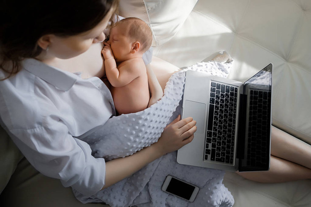 Cheerful young beautiful mother works as a freelancer at home, does remote work, breastfeeds her little child and reads information on a laptop. - Fotoğraf, Görsel