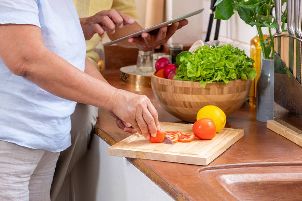 Aziatische Senior vrouw snijden tomaten op houten snijplank en het gebruik van Tablet PC om te zoeken menu recept. Senior met technologie lfiestyle. veroudering thuis - Foto, afbeelding