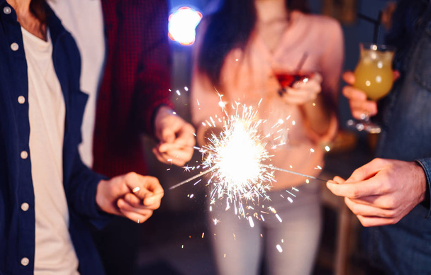 Group of happy people holding sparklers at party and smiling.Young people celebrating New Year together. Friends lit sparklers. Friends enjoying with sparklers in evening. Blur Background. - Zdjęcie, obraz