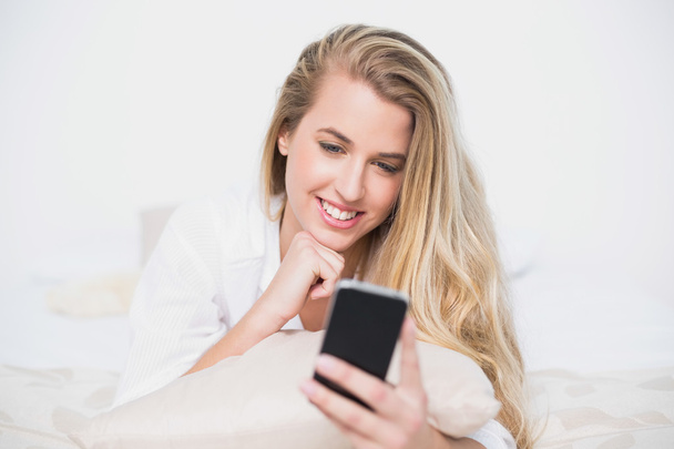 Smiling gorgeous model looking at her smartphone lying on cosy bed - Photo, image