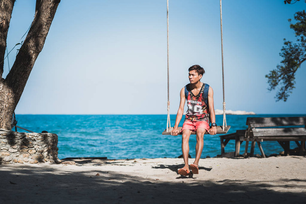 Le jeune homme s'assied sur une corde balançant sur le rivage sablonneux contre la mer bleue et le ciel clair. Homme asiatique porte un T-shirt avec "Swinging", "69" écrit dessus, tongs, dos & montre sur son bras. Ko Samet, Thaïlande
. - Photo, image
