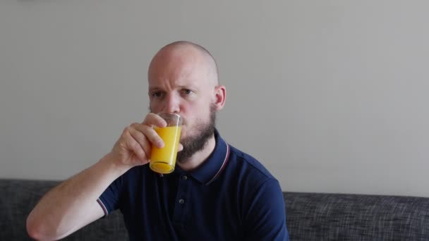 Knappe man met baard die vers sinaasappelsap drinkt en walgt van pulp. Grappig gezicht, marineblauw shirt, zittend op de slaapbank. - Video