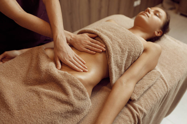 Female masseur pampering the stomach to young woman in towel, professional massage. Massaging and relaxation, body and skin care. Attractive lady in spa salon - Photo, Image