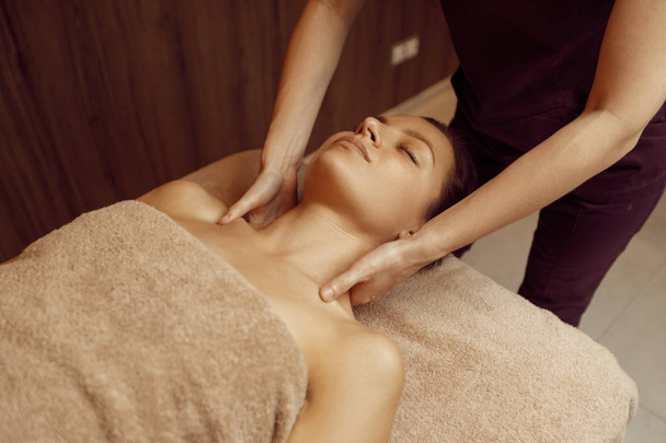 Female masseur pampering neck to young slim woman in towel, professional massage. Massaging and relaxation, body and skin care. Attractive lady in spa salon - Φωτογραφία, εικόνα