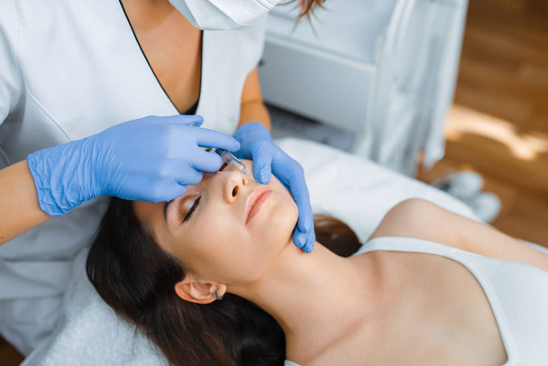 Cosmetician in gloves gives face botox injections to female patient on treatment table. Rejuvenation procedure in beautician salon. Doctor and woman, cosmetic surgery against wrinkles - Fotó, kép