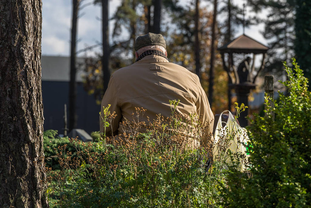 Un vieil homme gris vêtu d'un manteau et d'un chapeau clairs pleure sur sa tombe qui passe dans un cimetière à l'automne par une journée ensoleillée. Un panier placé à côté d'un homme
.  - Photo, image