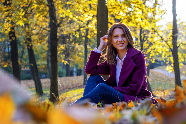 Όμορφη νεαρή γυναίκα με μπλε τζιν, λευκό πουκάμισο και μωβ παλτό κάθεται σε πεσμένα κίτρινα φύλλα στο πάρκο του φθινοπώρου. Κάτω όψη. - Φωτογραφία, εικόνα