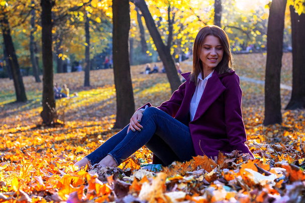 Дівчина в білій сорочці та фіолетовому пальто сидить на жовтому листі в осінньому парку. Релаксація в листопаді. Жовтневого вечора. Конгрес-журнал, спалахи
. - Фото, зображення