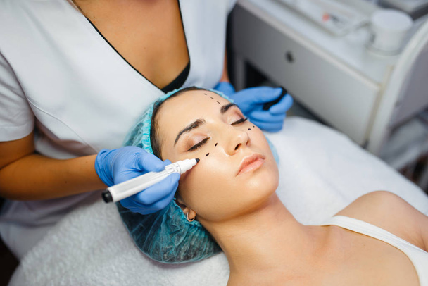 Cosmetician with marker puts dotted lines on female patient face, botox injections preparation. Rejuvenation procedure in beautician salon. Cosmetic surgery against wrinkles and aging - Photo, Image