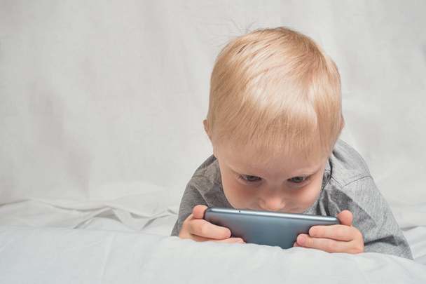 Niño rubio mirando algo su nariz enterrada en un teléfono inteligente, acostado en la cama. Gadget Ocio
 - Foto, imagen