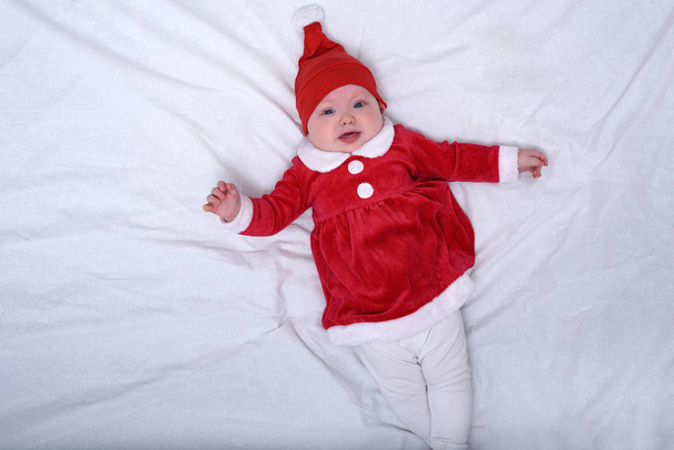 Portrét nespokojeného miminka v Santově klobouku a kostýmu. Vánoční nápad. Bílá postel. - Fotografie, Obrázek