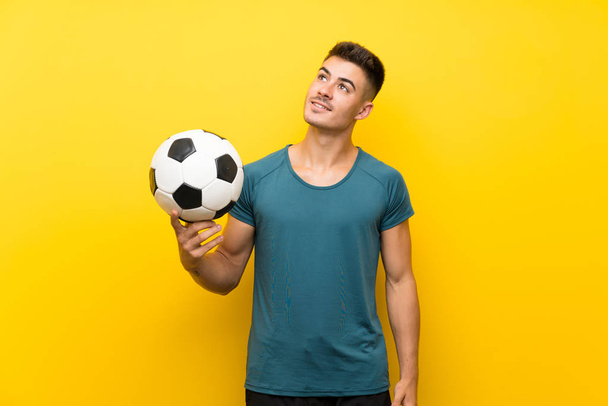 Przystojny młody piłkarz człowiek na odizolowanym żółtym tle patrząc w górę podczas uśmiechu - Zdjęcie, obraz