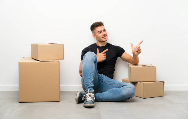 Handsome young man moving in new home among boxes pointing finger to the side - Foto, imagen