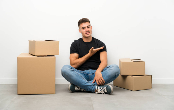 Handsome young man moving in new home among boxes extending hands to the side for inviting to come - Photo, Image