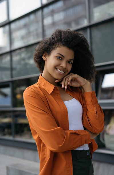 Portrait of young cheerful African American woman posing for pictures, standing outdoors, laughing, natural beauty. Hipster girl with beautiful smiling face, curly hairstyle walking on the street - Фото, изображение