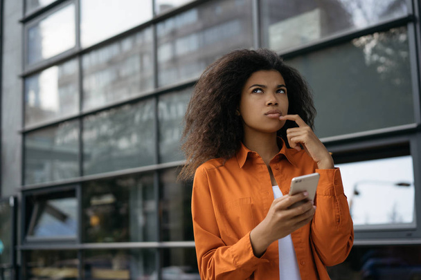 Pensive woman holding smartphone, shopping online, ordering food, booking tickets, searching information on website.  Young beautiful African American girl using mobile phone, waiting for taxi   - 写真・画像