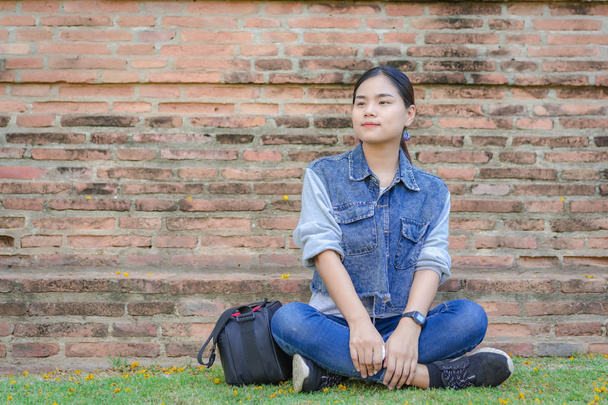 Portrait de jeune jolie femme asiatique relaxant et pensant plan d'avenir pour voyager. Une fille heureuse assise sur le terrain dans l'herbe verte dans un parc extérieur avec de vieux murs de briques arrière-plan
.   - Photo, image