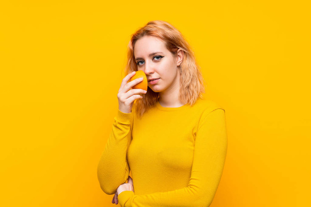 Young woman holding an orange over isolated yellow background - Фото, изображение