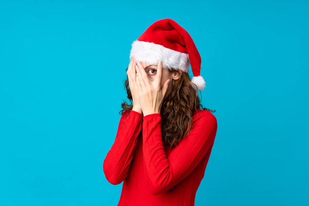 Girl with christmas hat over isolated yellow background covering eyes and looking through fingers - Фото, изображение