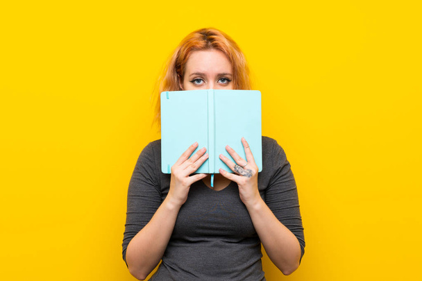 Young woman over isolated yellow background holding and reading a book - Fotoğraf, Görsel