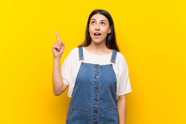 Young woman in dungarees over isolated yellow background pointing up and surprised - 写真・画像