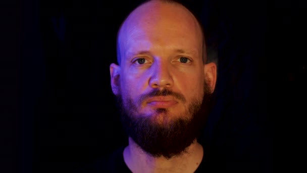 Bizar gevoel: vreemde man met baard die naar de camera staart en dan glimlacht, blauw en geel zacht licht op zwarte achtergrond - Video