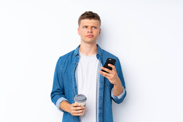 Νεαρός όμορφος άντρας πάνω από απομονωμένο λευκό φόντο κρατώντας καφέ για να πάρει μακριά και ένα κινητό ενώ σκέφτεται κάτι - Φωτογραφία, εικόνα