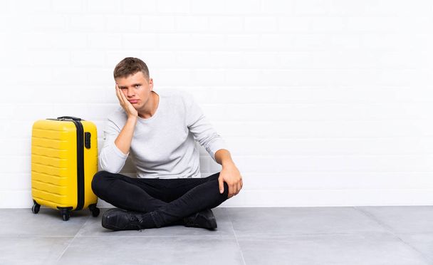 junger gutaussehender Mann sitzt mit einem Koffer unglücklich und frustriert auf dem Boden - Foto, Bild