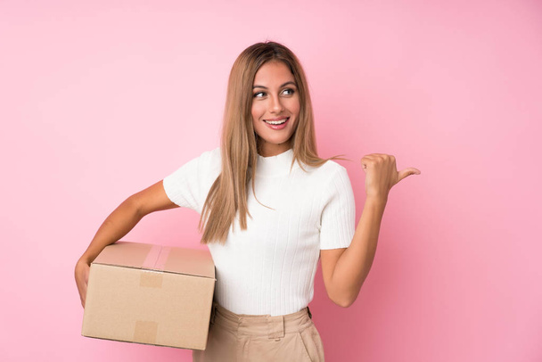 Young blonde woman over isolated pink background holding a box to move it to another site and pointing side - Φωτογραφία, εικόνα
