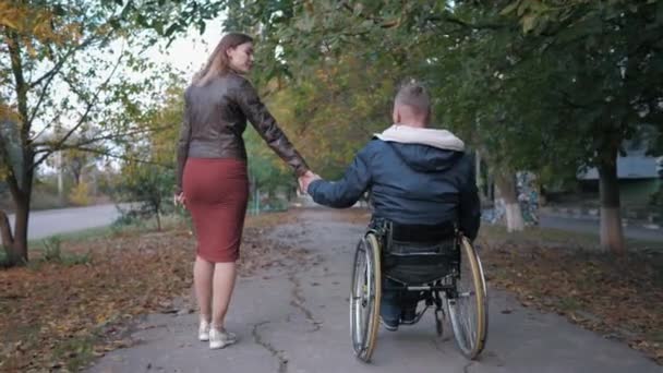 milovaná zdravotně postižená osoba na invalidním vozíku drží své ženy za ruku při chůzi v podzimním parku uprostřed stromů - Záběry, video