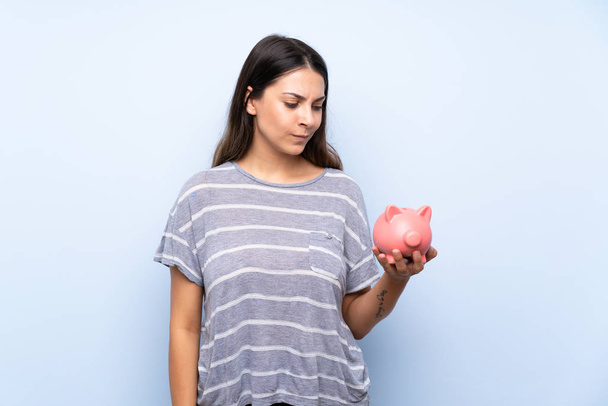 Young brunette woman over isolated blue background holding a big piggybank - Photo, Image