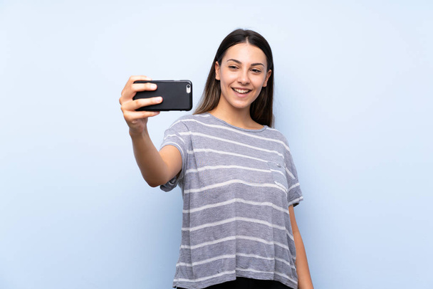 Young brunette woman over isolated blue background making selfie with cellphone - Valokuva, kuva