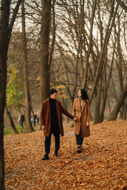 Stylish young couple outdoors on a beautiful autumn day in the forest. Young couple in love holding hands and walking through a park on a autumn day. The concept of youth, love and lifestyle. - Fotó, kép