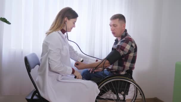 zdraví zdravotně postižené osoby, muž na invalidním vozíku je vyšetřen lékařem, měří tlak pomocí přístroje během lékařské prohlídky - Záběry, video