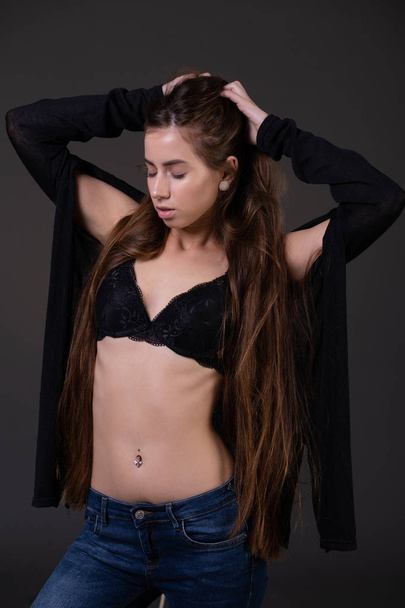 A beautiful young slender woman in a bra straightens her long hair closing her eyes posing on a black background - Zdjęcie, obraz