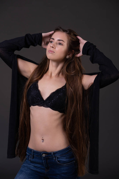 A beautiful young slender woman in a bra straightens her long hair, dressed in an unbuttoned jacket and jeans posing on a black background - Foto, imagen