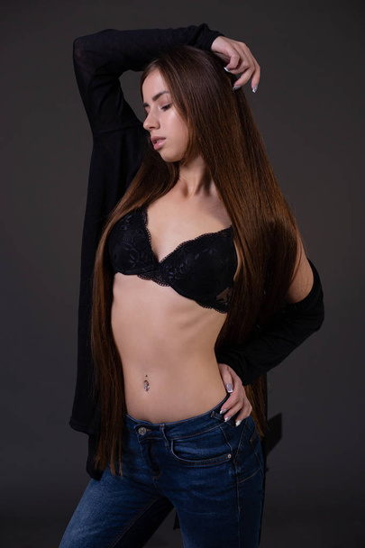A beautiful sexy young brown-haired woman dressed in jeans, a bra and an unbuttoned black jacket looks down thoughtfully - Foto, immagini