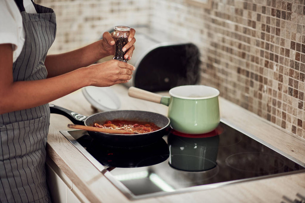Mulher de raça mista em avental de pé ao lado do fogão e adicionando pimenta em molho de tomate
. - Foto, Imagem