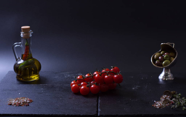 red tomatoes lie near a jug of oil, olives and spices on a dark background - Photo, Image