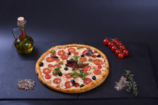 Italian pizza with tomatoes, peppers and meat lies next to a jug of spices on a dark background - Photo, image