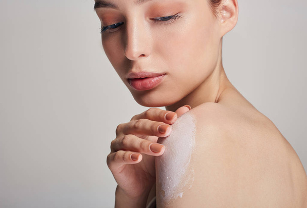 A young beautiful girl with brown hair and clean skin smears a moisturizer on the shoulder. Moisturizing and body care. - Foto, Bild
