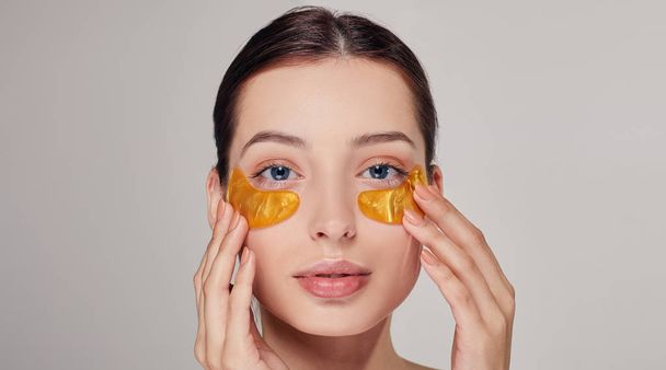 Young girl on the background applying golden collagen patches under the eyes. Mask for removing wrinkles and dark circles. - 写真・画像