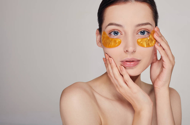 Pritty girl on the background applying golden collagen patches under the eyes. Mask for removing wrinkles and dark circles. A woman takes care of delicate skin around her eyes. Cosmetic procedures. - Foto, immagini