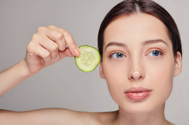 Beautiful attractive young lady with brown hair with full lips and full calm. The girl holds in her hand a slice of cucumber near her face. Looking side. Close up. - Foto, imagen