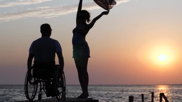 Nuori naaras seisoo laiturilla ja heiluttaa kangasta miespuolisen invalidin edessä pyörätuoliin iltahehkun taustalla valtameren yllä
 - Materiaali, video