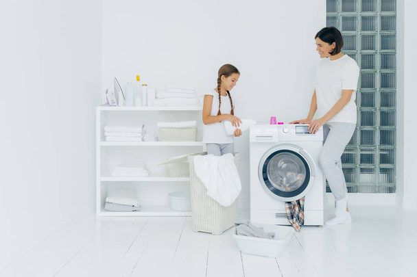 Критий знімок щасливої матері і дочки стоїть біля пральної машини, дівчина п'є рідкий порошок, завантажує пральну машину брудним одягом, виконує домашню роботу, прання вдома. Концепція домашніх справ
 - Фото, зображення