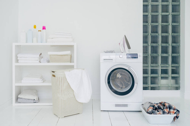 Laundry room with washing machine, dirty clothes in basket, iron and little shelf with neatly folded linen. Domestic room interior. Washing concept - 写真・画像