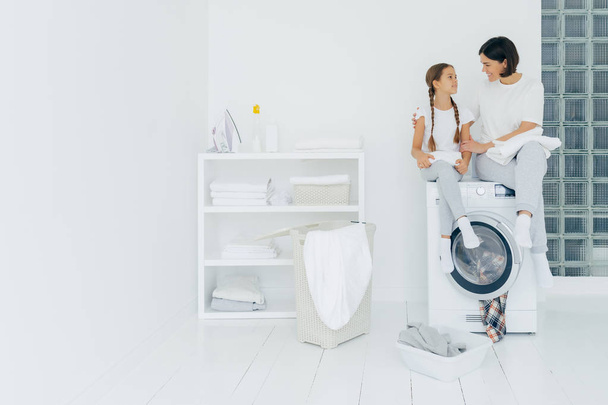 Happy caring woman embraces her little daughter, sit on washing machine, have rest after washing, surrounded with basket and dirty clothes, have pleasant conversation with each other. Housework - Photo, Image
