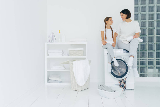Housewife and small child sit on top of washing machine, holds freshly washed towels, do washing together, wear white t shirts, pants and socks, white console with iron, folded linen and detergents - Photo, Image